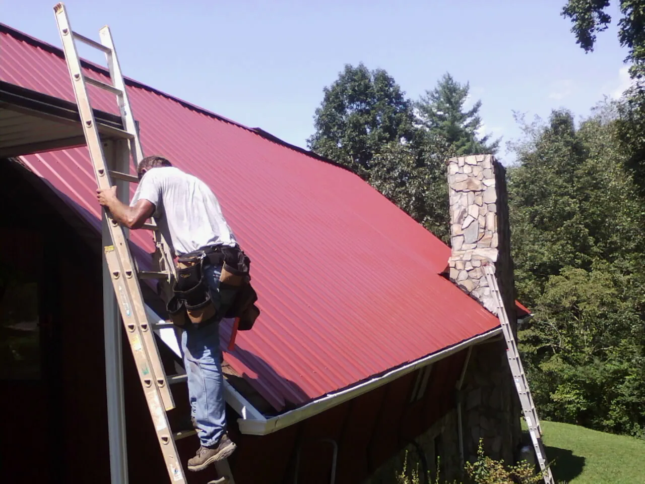 Roofer on a ladder beside a red metal roof and stone chimney — Soffit & Fascia Repair by Roofing Brothers in Estillfork